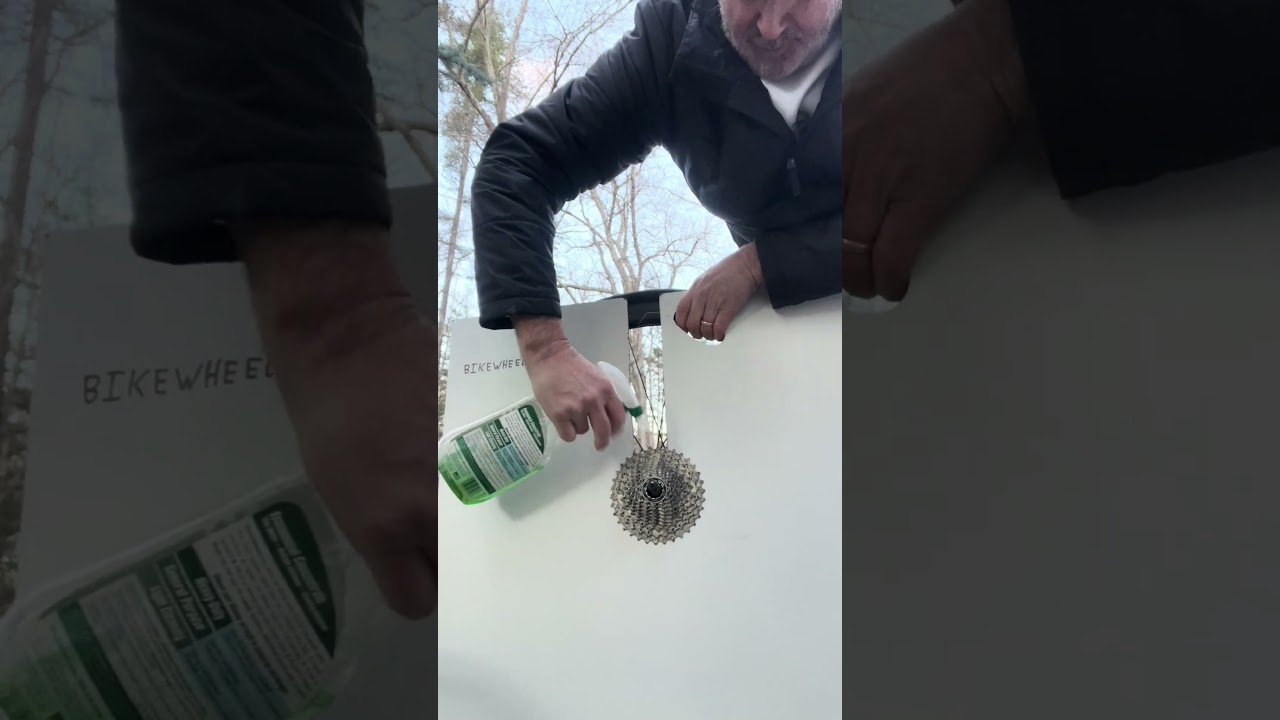 A person sprays cleaning solution on a bike cassette mounted against a white surface, demonstrating bike maintenance outdoors.